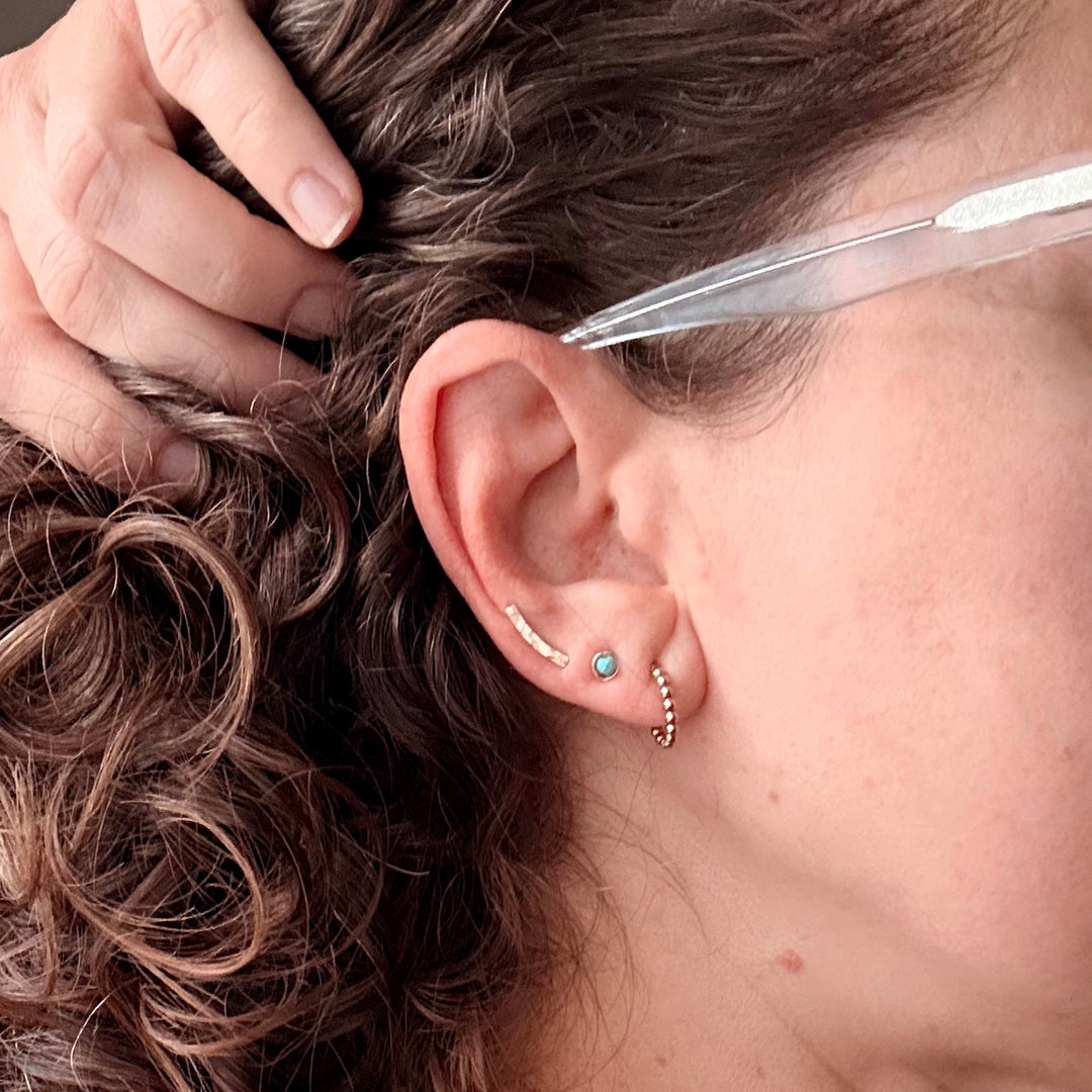 close up of ear wearing beaded ear huggers, birthstone studs with turquoise gemstones and 14k gold filled mini ear climbers